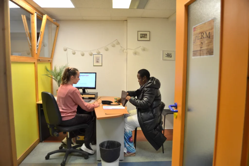 Deux femmes assises face à face à un bureau lors d'un entretien ; l'une travaille sur un ordinateur tandis que l'autre cherche dans son sac à main.